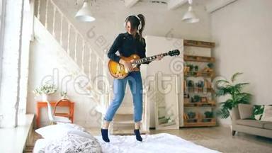 有趣的年轻女子戴着耳机在床上<strong>蹦蹦</strong>跳跳，家里的卧室里放着电吉他